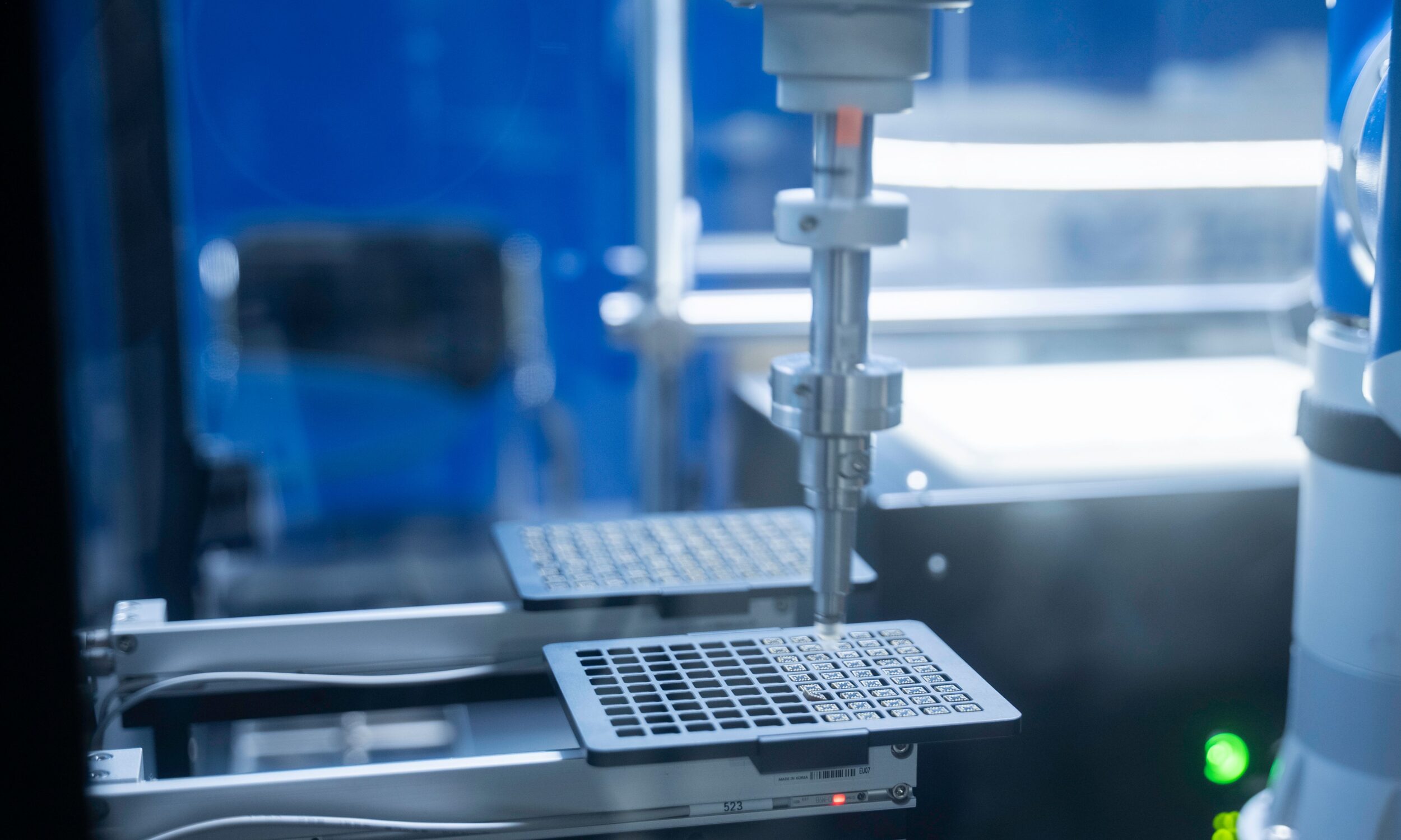 En robotarm placerer prøver i en bakke med rasterinddeling inde i et laboratorium, hvilket tyder på automatiseret udstyr til videnskabelige eller medicinske undersøgelser.
